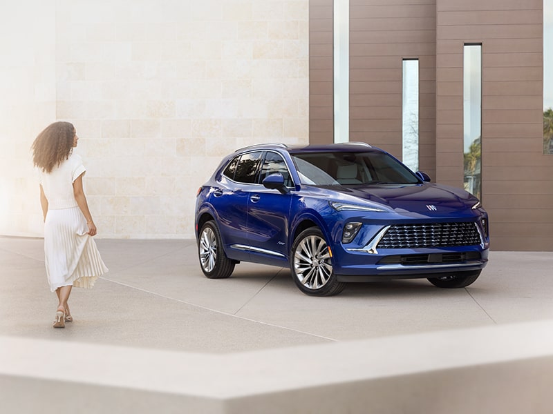 Wide Front Three-Quarters View of the 2025 Buick Envision Avenir Luxury SUV Near a Concrete Building with a Woman Walking Towards the Vehicle