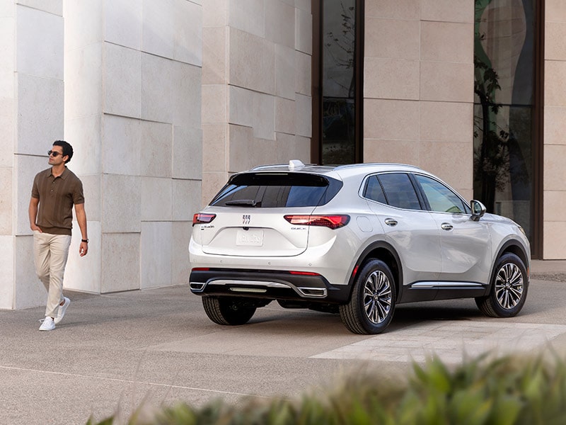 Wide Rear Three-Quarters View of a 2025 Buick Envision Small SUV Parked Near a Building with a Person Walking Away from the Vehicle