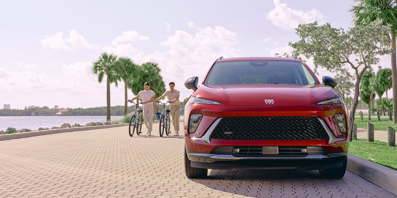 Front View of a Red Buick Envision Compact SUV Parked on a Tropical Street with Two Bikers Passing by